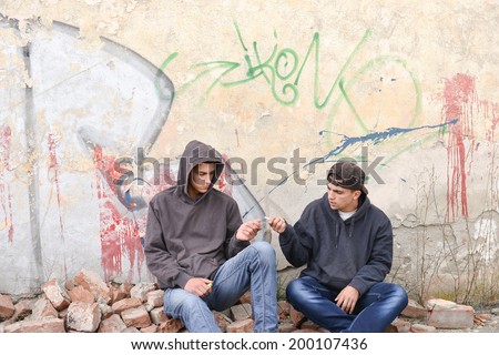 two street hooligans or rappers standing against a graffiti painted wall and sharing a cigarette