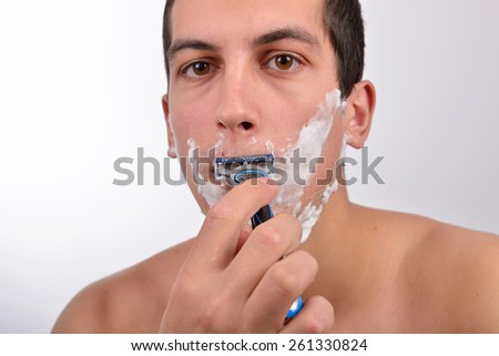 Handsome young man with lots of shaving cream on his face is shaving with razor