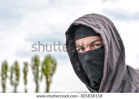 unrecognizable young man wearing black balaclava sitting on old stairs, looking at the camera