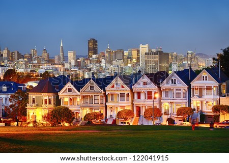 The Painted Ladies Of San Francisco, California Sit Glowing Amid The Backdrop Of A Sunset And Skyscrapers.