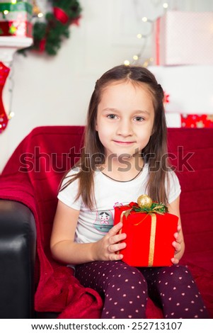 Little girl sitting on the couch holding a red box, Christmas gift, Christmas tree in the background