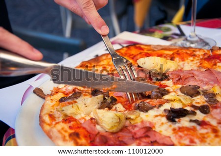 Cutting a delicious Italian pizza with cheese, mushrooms, artichokes and tomatoes with help of knife and fork. Main focus on fork