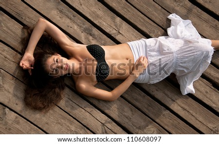 Pretty happy brunette girl in black bra with stresses and white long skirt with natural makeup lying on wooden planks outdoors