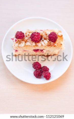 Top view on delicious sponge cake with raspberries and curds as filling with separate berries on a white plate with spoon on a textured table. Main focus on top raspberries on the cake