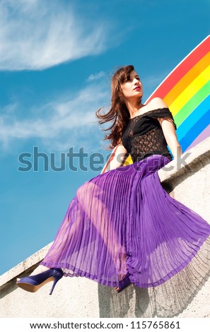 Bright colorful portrait of young brunette female model wearing bright purple skirt, black lace top and violet shoes sitting on wall against rainbow and bright blue sky background