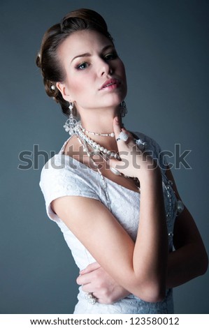 Low key studio shot of pretty bride wearing white dress,beautiful makeup, jewelry beads, pearls, earrings, rings, holding her hand near her neck, her lips open and looking at camera. Gray background