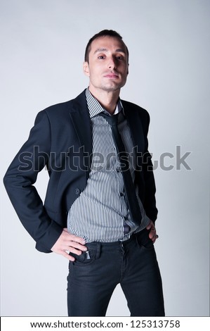 Confident successful stylish business man wearing black suit, stripped shirt, tie, hands on waist, standing and looking at camera from above with power and challenge. Gray background, copy space