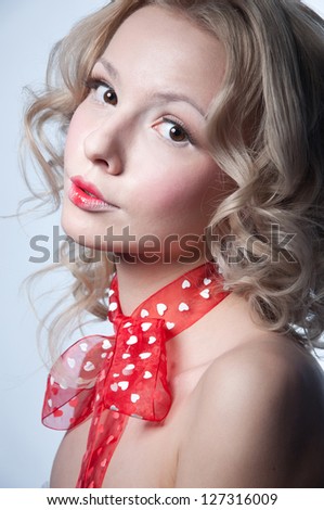 Pretty blond girl angel with curly hair, hazel eyes, wearing light makeup, red lips, red transparent ribbon with hearts made into a bow, shoulders naked, looking at camera. Copy space, gray background