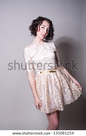 Studio portrait on gray of beautiful brunette woman model with curly hair, tender makeup wearing light golden dress with narrow belt, beige high heels, holding her dress, smiling, looking at camera