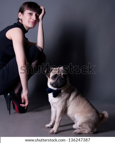 Studio portrait of an elegant pug dog gentleman wearing blue bow and sitting on the floor, with his owner beautiful young woman model sitting behind. Gray background