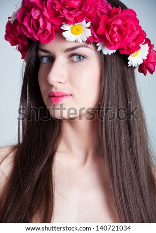 Closeup beauty studio portrait of young pretty woman with natural makeup, bright pink lips, wearing flower wreath made of red roses and chamomiles, looking at camera