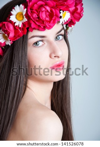 Closeup beauty studio portrait of young pretty woman with natural makeup, bright pink lips, wearing flower wreath made of red roses and chamomiles, looking at camera. Gray background, copy space