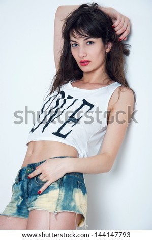 Pretty sexy woman model with great body, leaning on the wall, holding her arm behind head, wearing red lipstick, red nails, blue denim shorts, white top with black print, looking at camera. Copy space
