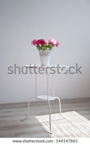 Pot with colorful red and pink rose flowers standing on a white table in minimalistic style in an empty room against the wall. Natural light from the window