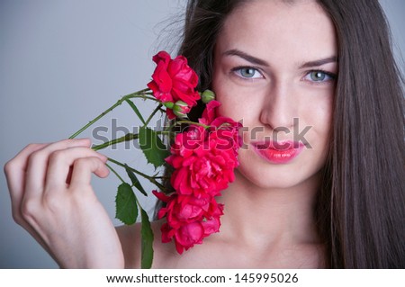 Young pretty woman model with healthy long straight brunette hair, natural makeup, tender glossy lips, gray eyes, holding twig with red roses near face, looking at camera and smiling. Grey background