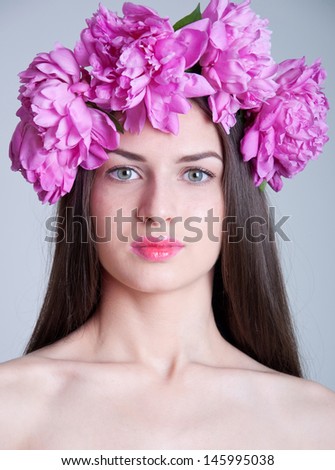Beauty studio portrait of young pretty woman with natural makeup, bright pink lips, wearing flower wreath made of big peonies, looking at camera. Grey background