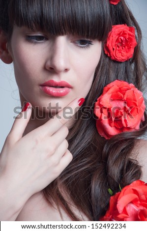 Pretty young woman model with red lips, elegant long brunette hair weaved into wave with big fresh pink terracotta roses, holding her hand near her chin, red nail polish. Looking down