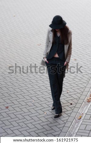 Pretty brunette woman model wearing stylish black pants suit, gray rain coat, black hat with wide fields, high heels, red lips, hands in pockets, going down the street covered with pavement tiles