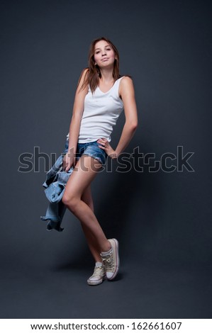 Cute and funny pretty young girl with very long legs wearing white top, blue denim shorts, sneakers, holding a jacket in her hands, standing, dancing and smiling. In studio over gray background