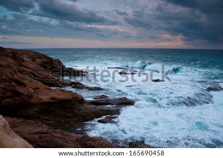 Stormy blue Ligurian sea with white spumy waves crashing against brown rocks. During sunset in autumn