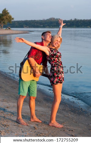 Young pretty couple of man and woman in love standing close to each other and dancing on the beach, man playing the guitar, both smiling, feeling happy together. During sunset