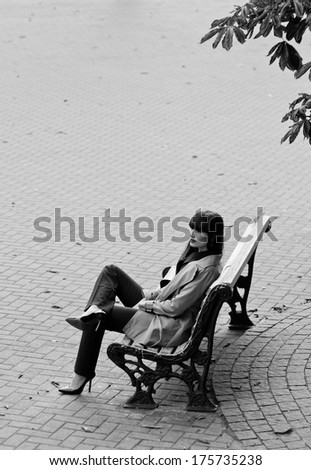Pretty brunette woman model wearing stylish black pants suit and gray rain coat, high heels, sitting on a bench with legs crossed high. In autumn on the street covered with pavement tiles. Copy space