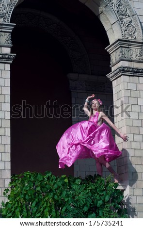 Barefooted beautiful young woman wearing flower wreath, silk airy dress, wind inflating this dress, standing on the porch covered with grape vines, against an old building with fretwork. Copy space
