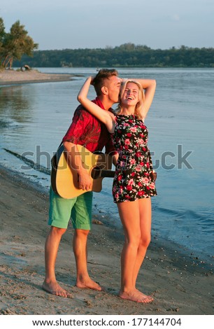 Young pretty couple of man and woman in love standing, dancing, singing on the beach close to each other, man playing the guitar, both smiling, feeling happy together. During sunset