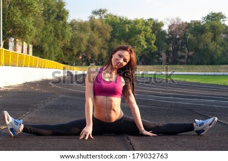 Pretty sporty strong slim and fit young woman sitting, doing split and stretching exercises, sitting on a full split. At stadium, on running tracks. During sunset. Green trees at background