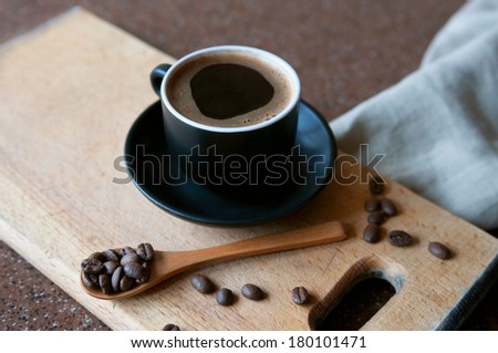 Still life with a hot espresso coffee with amazing cream in a black small cup on a saucer on a wooden board surrounded by roasted coffee beans in a wooden spoon. Copy space