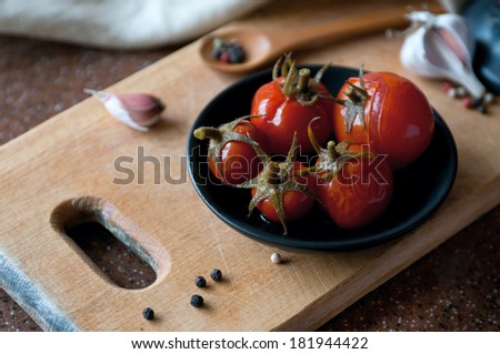 Kitchen still life of salted canned tomatoes on a saucer on a wooden board surrounded with a mortar and pestle, garlic cloves and red, white, black pepper. Main focus on the stem of tomatoes