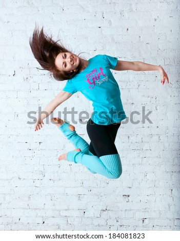 Pretty sporty barefooted woman dancer wearing black leggings, blue t-shirt and gaiters, dancing, jumping up in the air, posing, smiling and looking happily at camera. Over a brick wall background