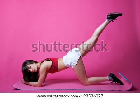 Pretty sporty woman trainer doing workout in studio - doing leg swings to make her butt look sexy. Over pink background, copy space