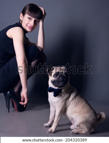 Studio portrait of an elegant pug dog gentleman wearing blue bow tie and sitting on the floor, with his owner beautiful young woman model sitting behind. Gray background, copy space
