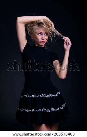 Blond woman model wearing black dress, holding her curly hair, looking at with surprise and disappointment. Over blackbackground