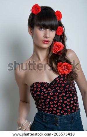 Pretty young woman model with red lips, elegant long brunette hair weaved into wave with big fresh terracotta roses, black corset underwear with red lips pattern, blue denim shorts. Looking at camera