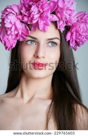 Beauty studio portrait of young pretty woman with natural makeup, bright pink lips, wearing flower wreath made of big peonies, looking at camera. Grey background