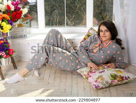 Cute funny teenage girl wearing pijamas, lying on the floor on pillows, enjoying her morning, smiling and looking at camera. Near the big french window