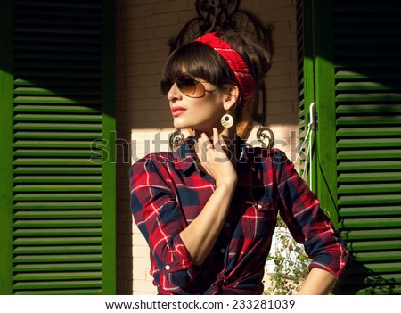 Pretty brunette woman wearing red and blue checked shirt, band, aviator sunglasses, resting in a street cafe, posing against the wall and green shutters. On a hot summer day