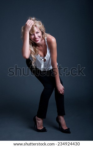 Pretty happy blond business woman wearing high heels, white top and black pants, red lipstick, hair made in locks, laughing, posing, looking at camera, smiling with toothy smile. Over gray background