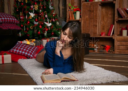 Pretty brunette woman wearing a warm knitted sweater, lying on a fur cover on the floor near decorated Christmas tree in a living room, reading a book. Many presents and pillows nearby.