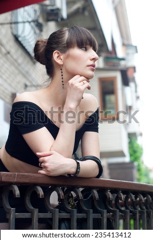 Pretty slim young brunette woman model wearing black off-the-shoulder top, elegant earrings, beautiful hairstyle, leaning over balustrade of a balcony, looking somewhere, dreaming and enjoying the day