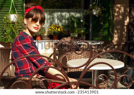 Pretty brunette woman wearing red skirt, checked shirt, band, aviator sunglasses, resting in a wickered chair near the table in a street cafe, looking at camera. Bright open light. On a hot summer day