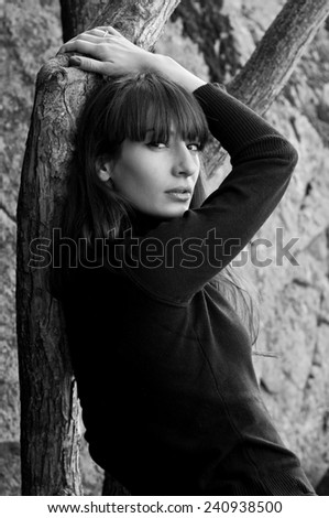 High fashion black and white  portrait of pretty stylish young woman outdoors wearing black turtle-neck, standing near a tree, leaning against it, against rocks background, looking at camera.