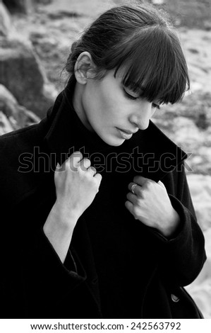 Black white portrait of elegant beautiful brunette woman model wearing a turtleneck sweater, holding her coat collar with hands, posing, looking down, near rocks on a bright sunny day. Autumn fashion.