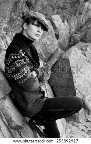 Black white portrait of elegant beautiful brunette woman model in boyish clothes like turtleneck sweater, knitted jacket, jeans, cap, red nails, patent leather shoes, siting near rocks. Autumn fashion