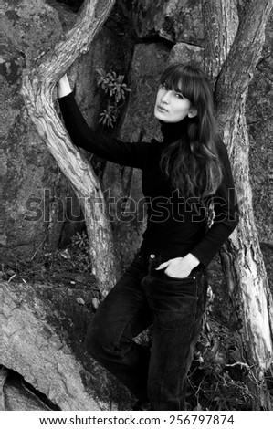 Pretty brunette woman model wearing corduroy pants, turtleneck polo, standing near trees and stone rocks, posing, looking at camera. Play of lines with tree and hair curves. Natural light, copy space