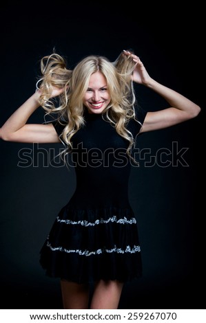 Pretty blond young emotional woman model with curly hair wearing black dress, red lipstick, holding her hair with hands, shouting, having fun. Over black background