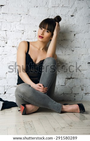 Pretty sexy woman model with amazing body long legs seating on floor wearing denim jeans black lingerie top high heels posing looking at camera jacket nearby. White brick wall background copy space