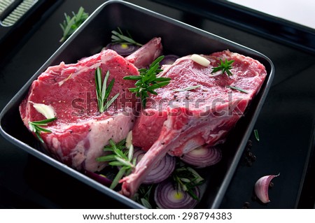 Raw fresh meat beef steak, seasonings with salt, pepper, rosemary leaves and red onion, on a black tray ready to be cooked